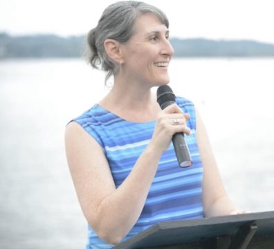 Dr. Heather Price speaking at a climate strike, in front of a lake in Kirkland, WA in 2019. She's wearing a dress that has blue and white stripes at the top, and orange and red strips at the bottom, representing the change in climate over the last century.