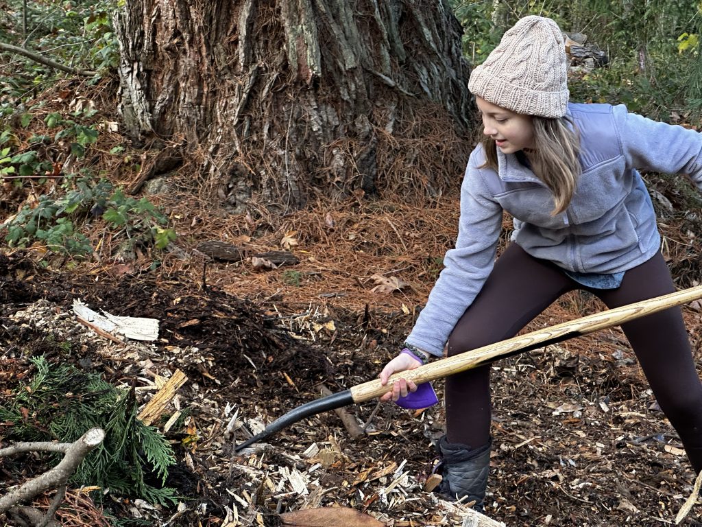 young person wearing a cream hat and purple jacket, using a pitchfork to shovel mulch